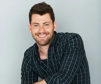 A man with a beard and plaid shirt smiles at the camera, against a plain gray background.