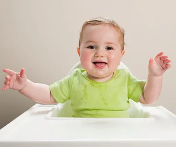 "Happy baby in a green shirt sitting in a white high chair, smiling and raising arms."