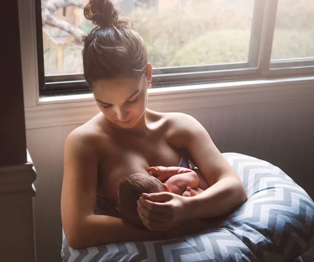 A mother breastfeeding her baby using a nursing pillow by a window.