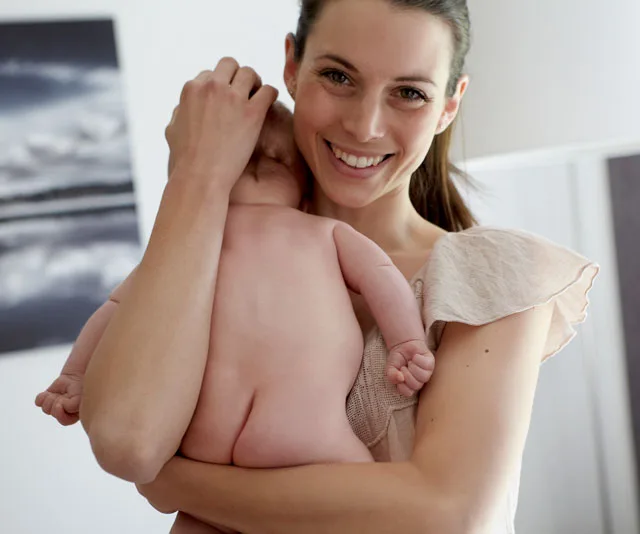 Mother smiling while holding a baby over her shoulder in a cozy home setting.