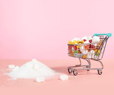 Mini shopping cart with sugar cubes and candies, next to a pile of sugar, on a pink background.