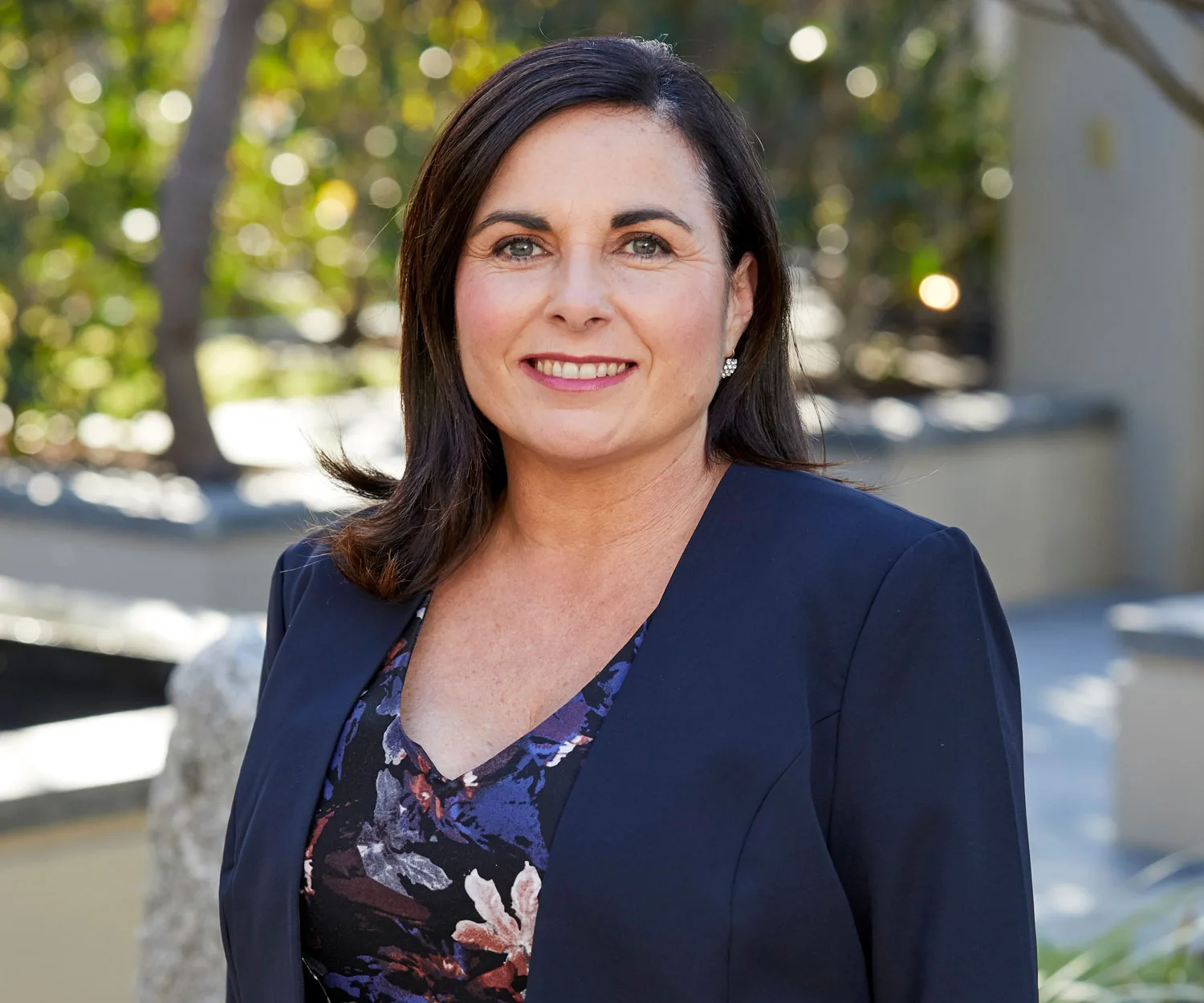 A woman with dark hair in a navy blazer smiles outdoors with a blurred background of greenery.
