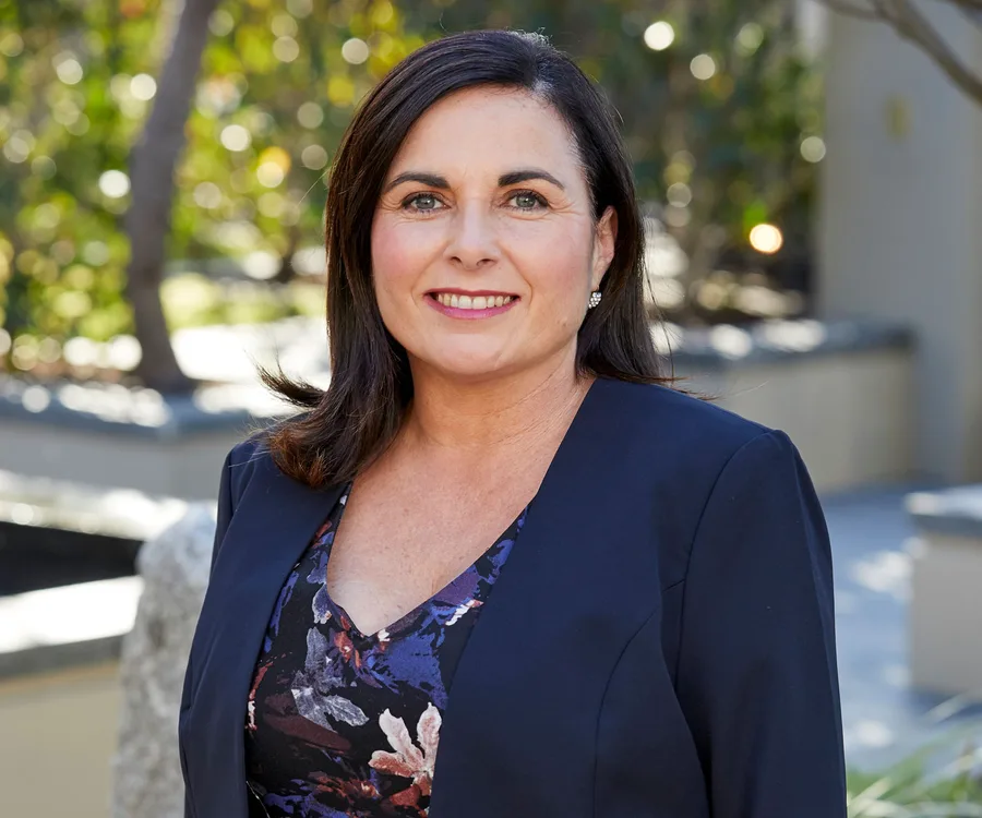 A woman with dark hair in a navy blazer smiles outdoors with a blurred background of greenery.