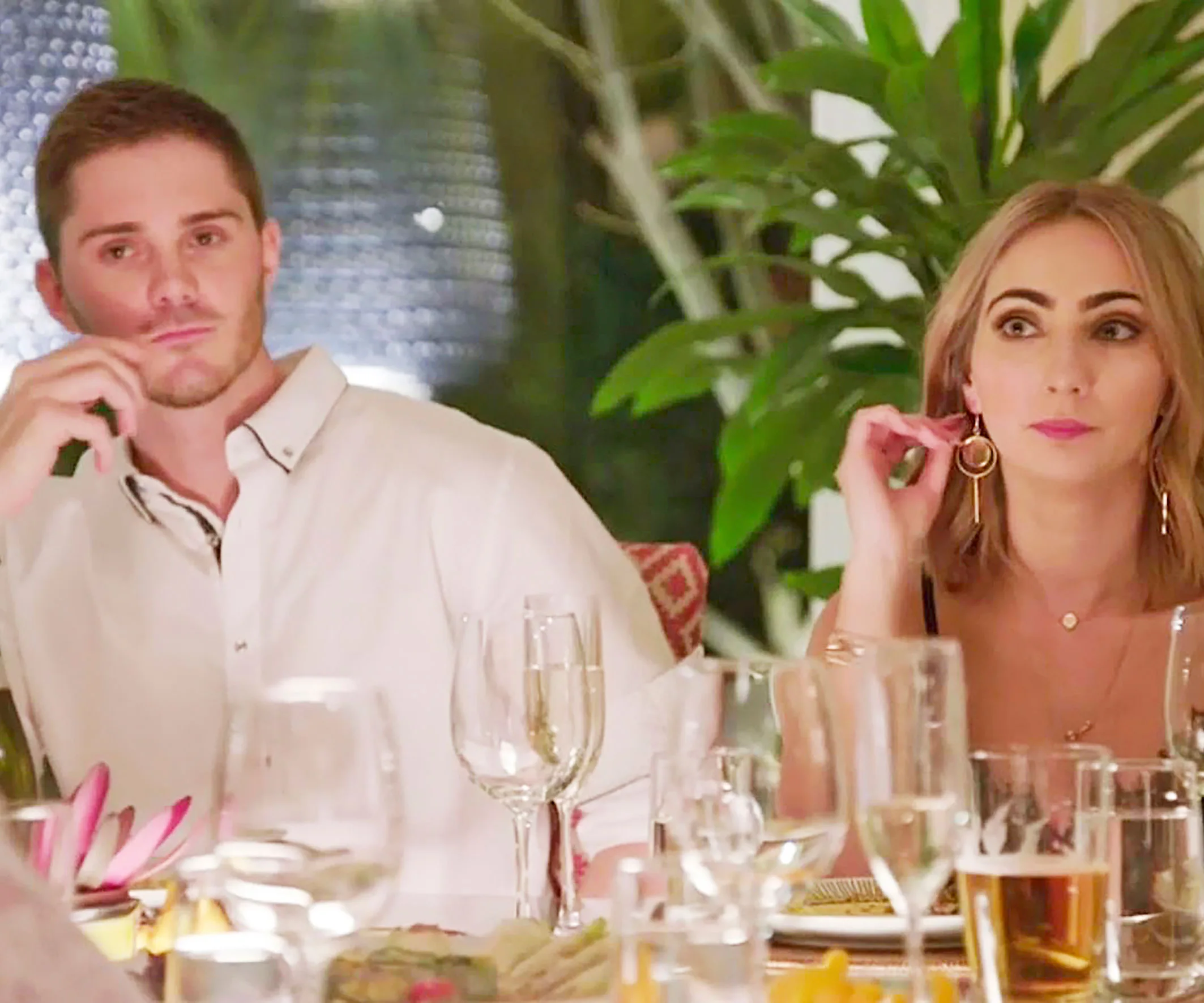 Young man and woman seated at a dinner table with drinks, plants in the background.