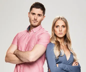 A man in a pink shirt and a woman in a blue top stand back-to-back, arms crossed, looking serious.