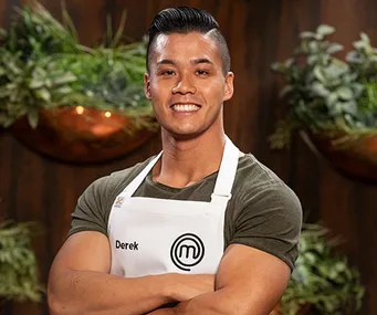 Derek Lau, a contestant from MasterChef, smiles confidently with arms crossed, wearing a white apron with the MasterChef logo.
