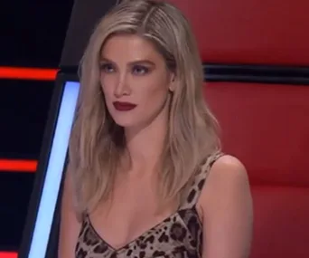 Delta Goodrem in a leopard print dress sits on a red chair during "The Voice" episode.