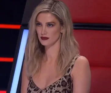 Delta Goodrem in a leopard print dress sits on a red chair during "The Voice" episode.