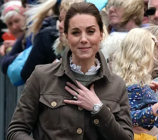 "Kate Middleton greets a crowd in Cumbria, wearing a brown jacket, with people and children in the background."