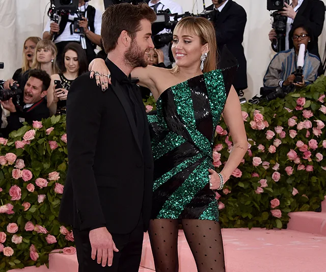 Couple smiling at each other on a pink carpet surrounded by photographers and floral decorations.