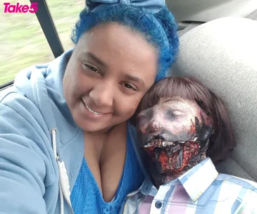 A person with blue hair smiles, hugging a life-like zombie doll with decayed features inside a car, "Take 5" logo visible.