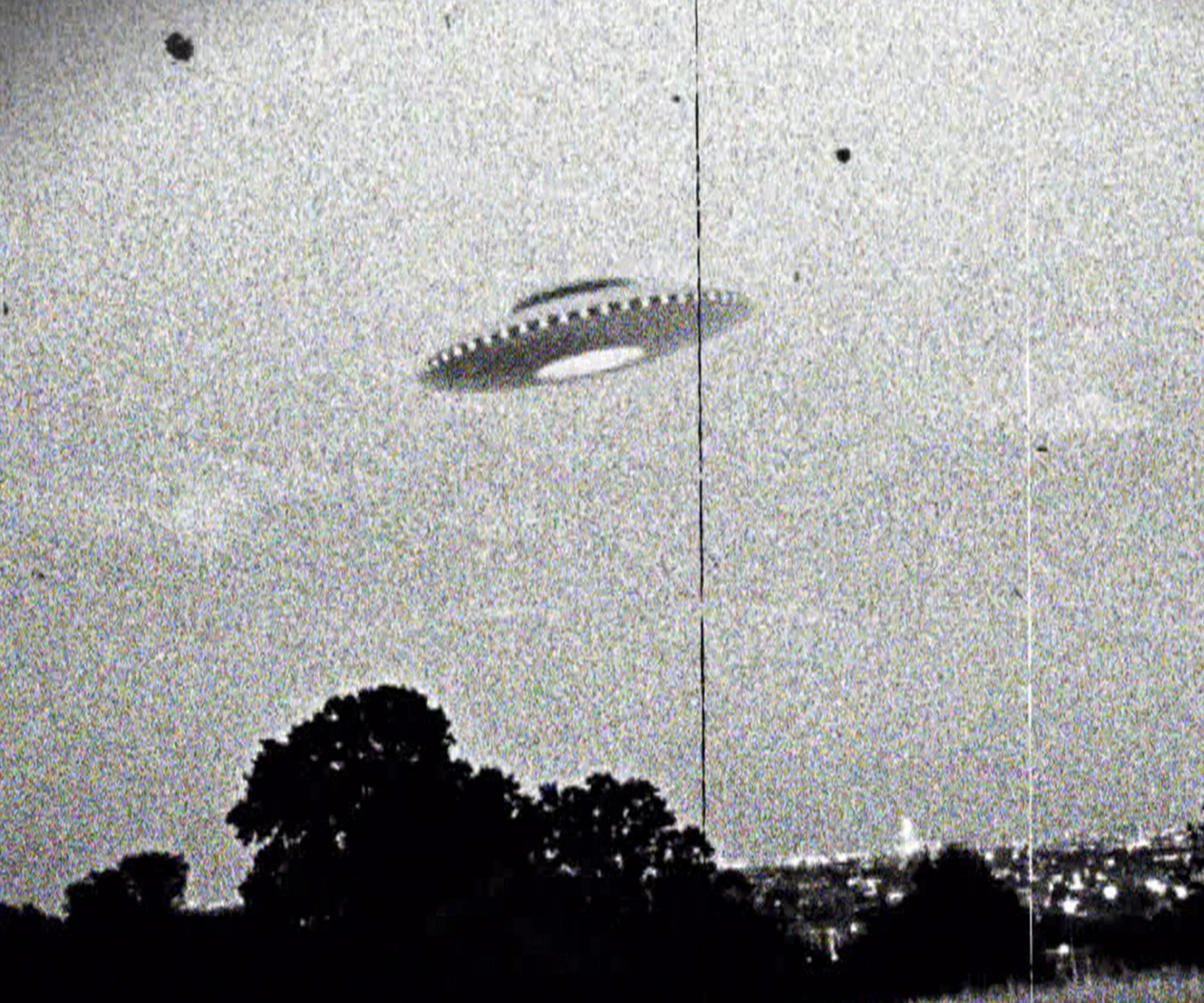 Grainy black-and-white photo of a UFO flying over a landscape with trees and distant city lights.