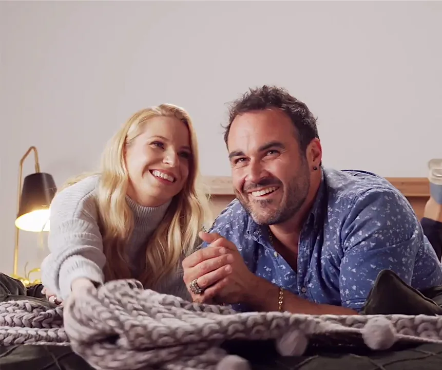 Happy couple lying on bed with a knitted blanket, smiling at something off-camera.