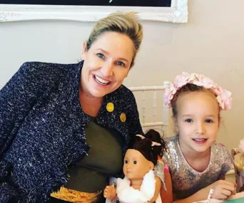 Smiling woman and young girl with a doll, seated at a table, girl wearing a flower headband.