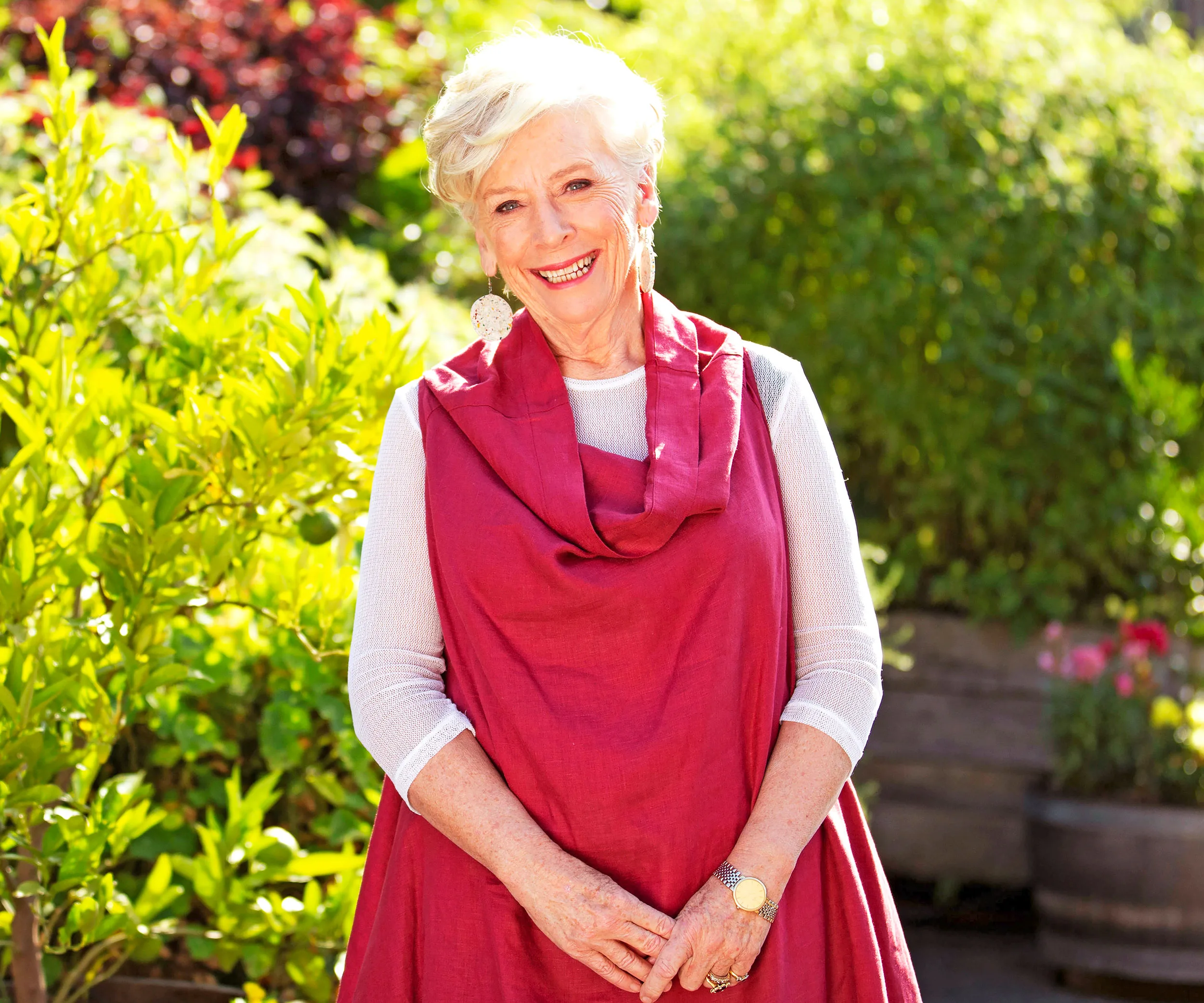 An elderly woman smiling in a garden, wearing a red shawl and white top, with bright sunlight and greenery around her.