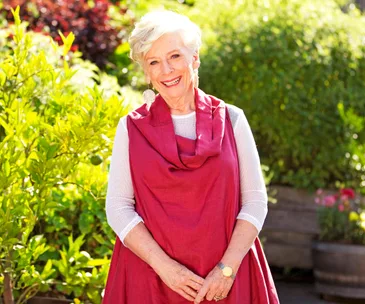 An elderly woman smiling in a garden, wearing a red shawl and white top, with bright sunlight and greenery around her.