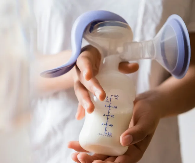 Hands holding a breast pump with milk inside, showing measurement markings on container.
