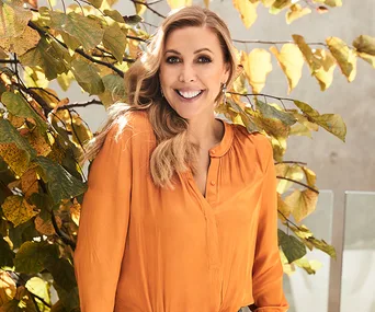 Woman in an orange top standing outdoors, smiling with autumn leaves in the background.