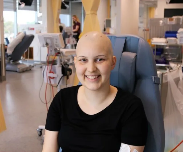 A smiling person with a shaved head sits in a hospital chair, suggesting a chemotherapy treatment setting.