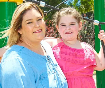 A woman and young girl smiling outdoors, with the girl wearing a pink dress on playground equipment.
