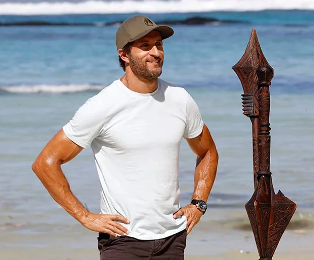 Man in a white t-shirt and cap stands on a beach with a carved tribal staff.
