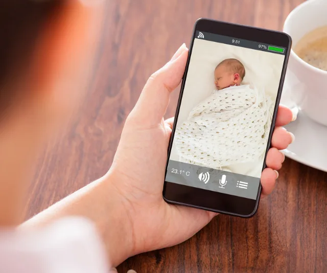 Person viewing a baby monitor app on a smartphone, showing a sleeping baby wrapped in a blanket.