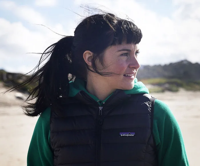 A person with a fringe and ponytail, wearing a vest and hoodie, stands on a beach, looking sideways.