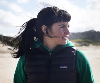 A person with a fringe and ponytail, wearing a vest and hoodie, stands on a beach, looking sideways.
