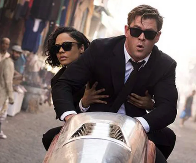 Agents in black suits wearing sunglasses ride a sleek, futuristic vehicle through a crowded market street.