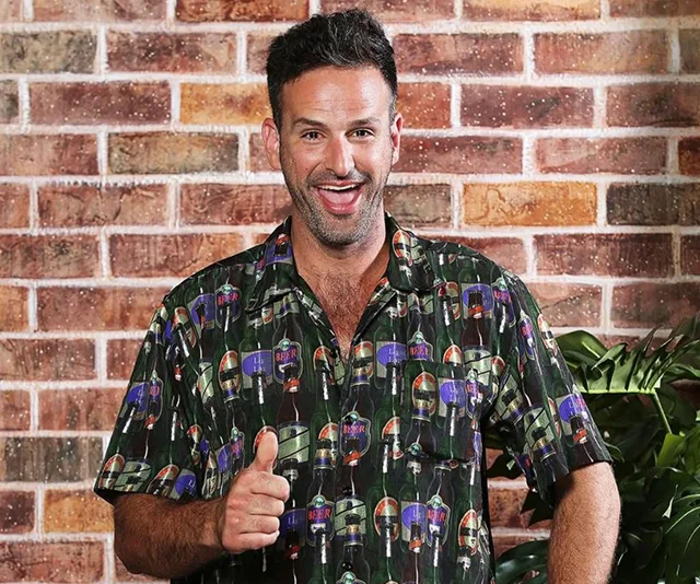 A person smiling and giving a thumbs-up, wearing a printed shirt, standing against a brick wall background.