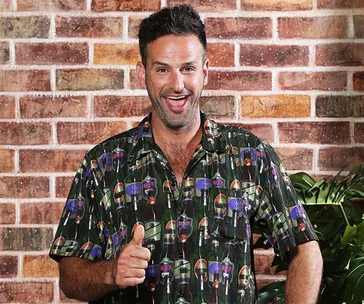 A person smiling and giving a thumbs-up, wearing a printed shirt, standing against a brick wall background.