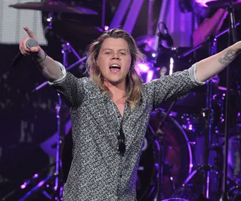 Singer performing on stage with arms raised, microphone in hand, in front of a drum set and purple lights.