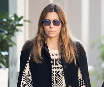Woman with long hair wearing sunglasses and a patterned sweater under a dark coat, standing outdoors near greenery.