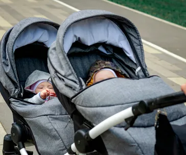 Two babies sleeping in a side-by-side double stroller on a paved path.