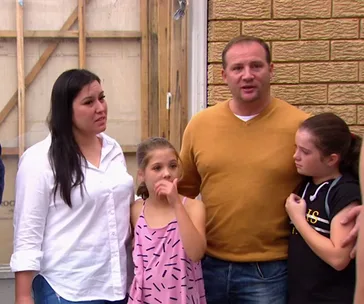 Family standing outside their house, with a man in a yellow sweater and a woman comforting two children.