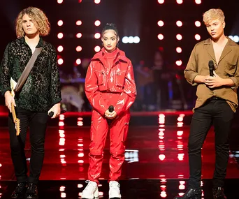 Three contestants stand on a stage with microphones during a performance show, surrounded by bright stage lights.