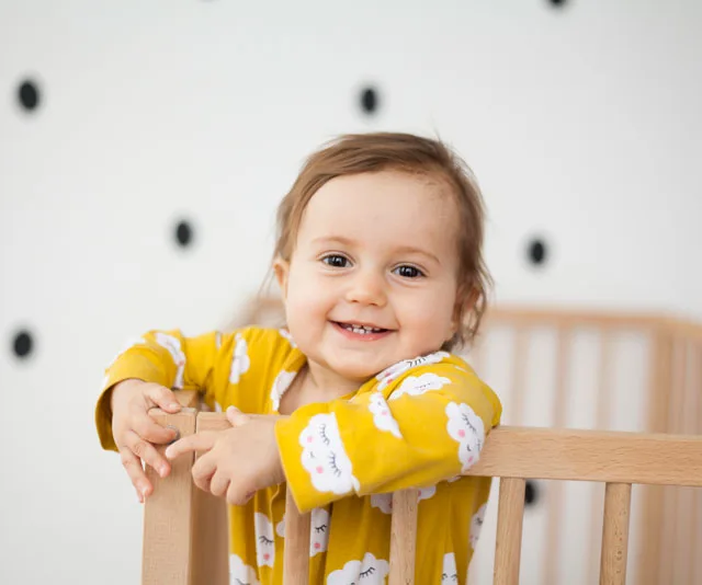A happy toddler in a yellow cloud-patterned outfit stands in a wooden crib, with a polka-dot wall in the background.