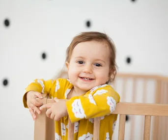 A happy toddler in a yellow cloud-patterned outfit stands in a wooden crib, with a polka-dot wall in the background.