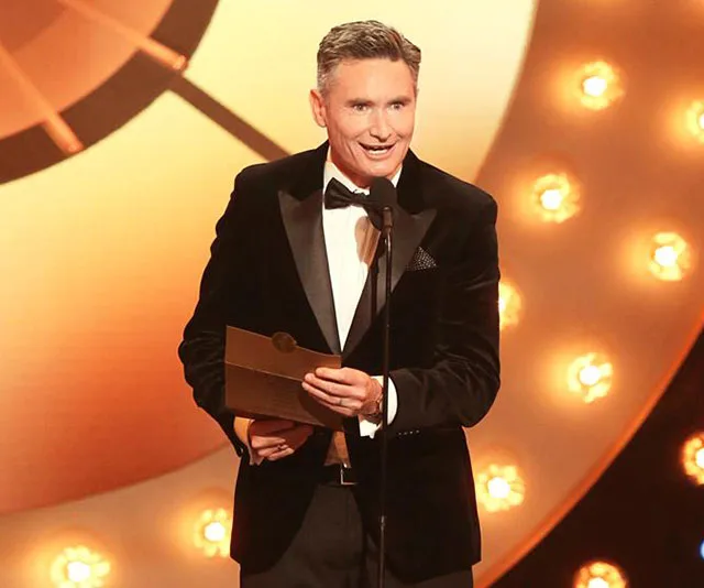 Man in a tuxedo speaks on stage, holding a brown envelope, with a bright circular backdrop and lights.