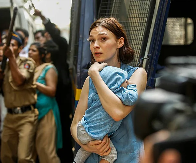 A worried woman holds a baby outside a police vehicle, surrounded by officers and onlookers in a crowded street scene.