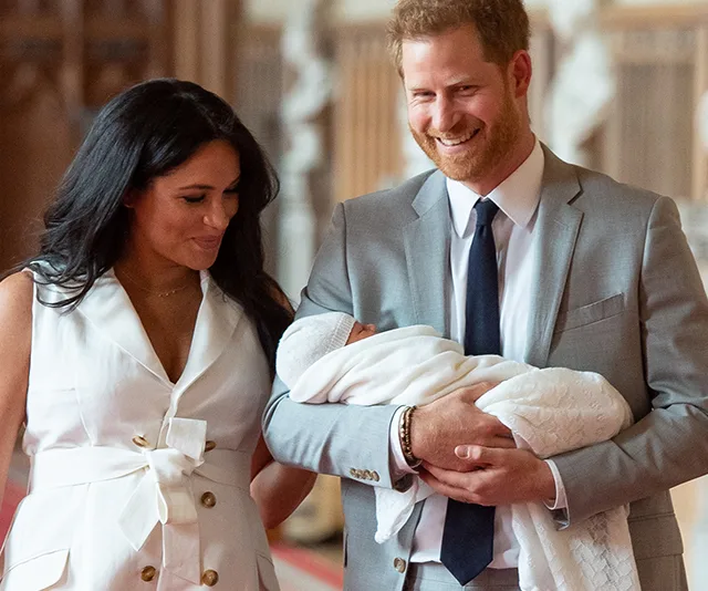 A couple smiling at each other, holding a baby wrapped in a white blanket at a formal event.