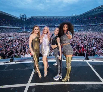 Four performers on stage at a large concert venue with a cheering crowd in the background.