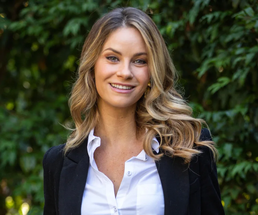 Smiling woman with long hair wearing a white shirt and black blazer, standing in front of green foliage.