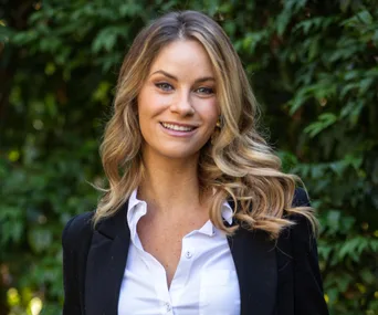 Smiling woman with long hair wearing a white shirt and black blazer, standing in front of green foliage.