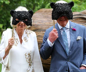 Couple in formal attire holding black sequin animal masks on sticks, standing outside with a large sculpture behind.