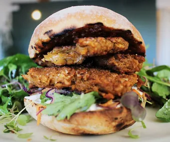 Vegan sandwich with plant-based patty, lettuce, and sauce, served on a white plate with greens in the background.