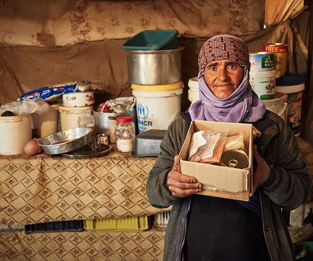 A woman holding a box of food supplies in a makeshift kitchen setting, with containers and items in the background.