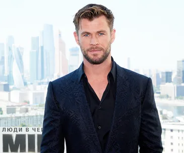 Actor in a blue suit standing with a cityscape background during a sunny day event.