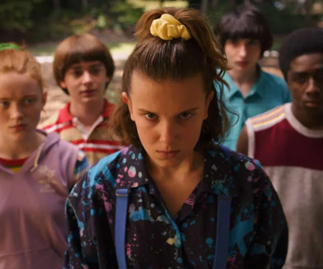 Five teens stand outdoors, focused and serious, with the girl in front wearing a scrunchie and patterned shirt.