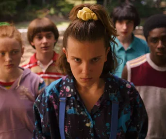 Five teens stand outdoors, focused and serious, with the girl in front wearing a scrunchie and patterned shirt.